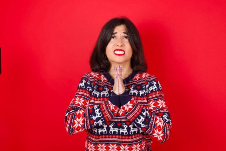 Photo for Brunette caucasian woman wearing christmas sweater over red background begging and praying with hands together with hope expression on face very emotional and worried. Please God - Royalty Free Image