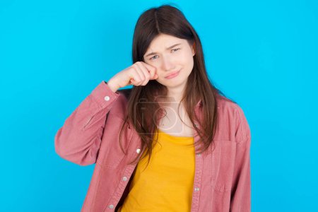 Téléchargez les photos : Malheureux jeune fille caucasienne portant chemise rose isolé sur fond bleu pleurer tout en posant à la caméra fouettant des larmes avec la main. - en image libre de droit