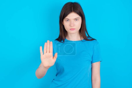 Photo for Young caucasian girl wearing blue T-shirt isolated over blue background shows stop sign prohibition symbol keeps palm forward to camera with strict expression - Royalty Free Image