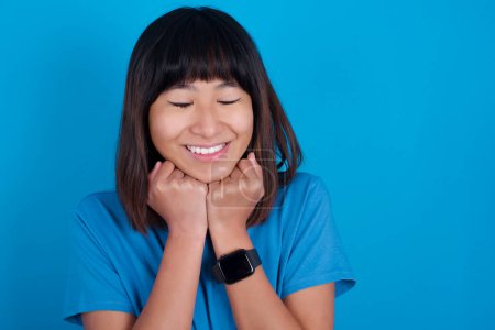 Téléchargez les photos : Joyeux jeune asiatique femme portant t-shirt sur fond bleu a timide expression satisfaite, sourit largement, montre dents blanches, Les gens émotions - en image libre de droit