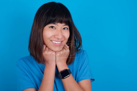 Téléchargez les photos : Satisfait jeune femme asiatique portant t-shirt sur fond bleu touche menton avec les deux mains, sourit agréablement, se réjouit bonne journée avec amant - en image libre de droit