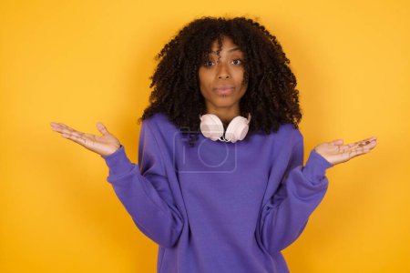 Foto de Retrato de joven mujer afroamericana expresiva con auriculares sobre hombros de trituración de fondo amarillo - Imagen libre de derechos