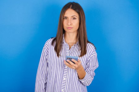 Photo for Upset dissatisfied beautiful young woman uses mobile software application and surrfs information in internet, holds modern mobile hand - Royalty Free Image