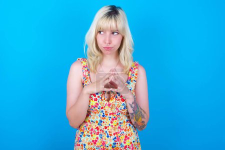 Photo for Caucasian girl wearing floral dress isolated over blue background steepls fingers and looks mysterious aside has great evil plan in mind - Royalty Free Image