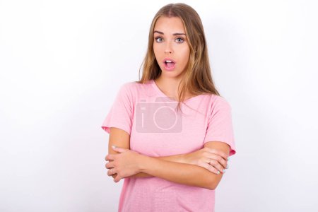 Téléchargez les photos : Choqué embarrassé Jeune fille caucasienne portant T-shirt rose sur fond blanc garde la bouche largement ouverte. Entend une nouveauté incroyable fixer dans la stupeur - en image libre de droit