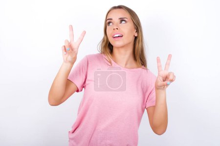 Photo for Isolated shot of cheerful Young Caucasian girl wearing pink T-shirt on white background makes peace or victory sign with both hands, feels cool. - Royalty Free Image