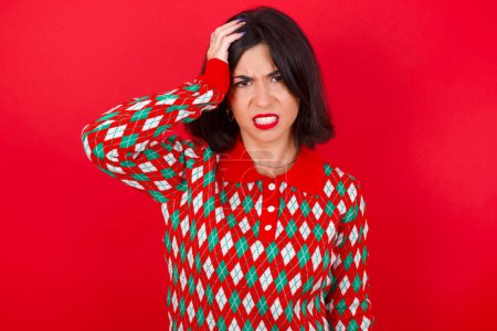 Téléchargez les photos : Embarrassé femme caucasienne brune portant chandail de Noël sur fond rouge avec une expression choquée, exprime grand étonnement, pose modèle perplexe intérieur - en image libre de droit