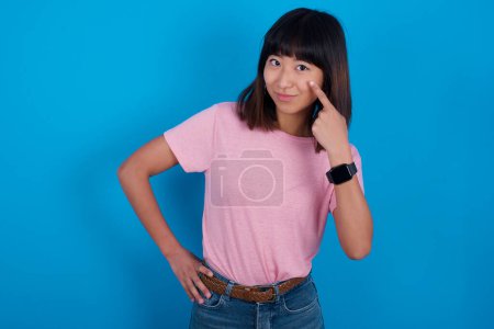 Téléchargez les photos : Jeune femme asiatique portant t-shirt sur fond bleu regardant, observant, gardant un oeil sur un objet devant, ou regarder dehors pour quelque chose. - en image libre de droit
