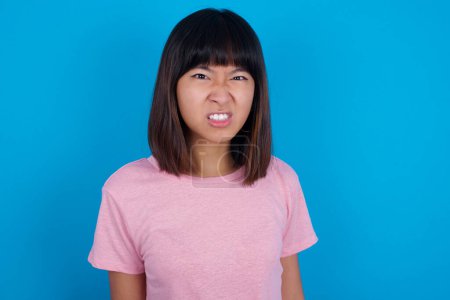 Photo for Mad crazy young asian woman wearing t-shirt against blue background clenches teeth angrily, being annoyed with coming noise. Negative feeling concept. - Royalty Free Image