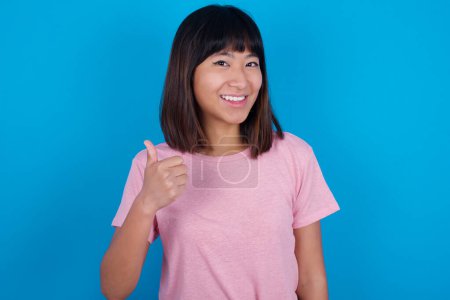 Photo for Young asian woman wearing t-shirt against blue background giving thumb up gesture, good Job! Positive human emotion facial expression body language. - Royalty Free Image