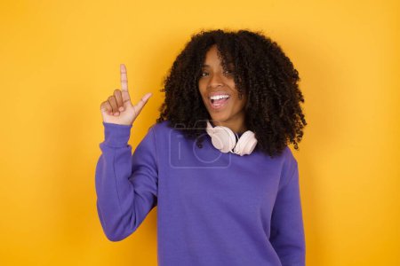 Photo for Portrait of young expressive african american woman with headphones on yellow background showing index finger - Royalty Free Image