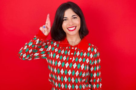 Téléchargez les photos : Brunette femme caucasienne portant chandail de Noël sur fond rouge pointant vers le haut avec les doigts numéro dix en chinois langue des signes Shi - en image libre de droit