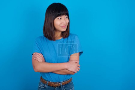 Téléchargez les photos : Agréable jeune femme asiatique portant t-shirt sur fond bleu garde les mains croisées sur la poitrine semble heureusement de côté - en image libre de droit