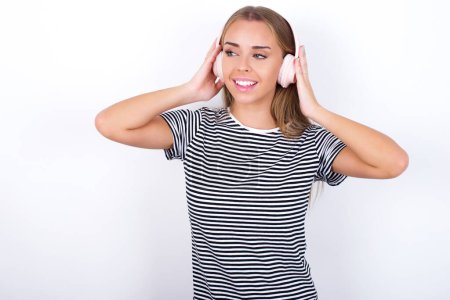 Photo for Beautiful blonde girl wearing striped t-shirt on white background wears stereo headphones listens music concentrated aside. People hobby lifestyle concept - Royalty Free Image