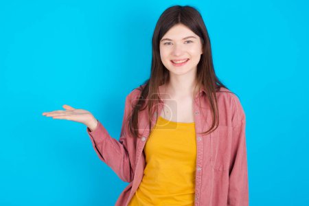 Photo for Positive glad young caucasian girl wearing pink shirt isolated over blue background says: wow how exciting it is, has amazed expression, shows something on blank space with open hand. Advertisement concept. - Royalty Free Image