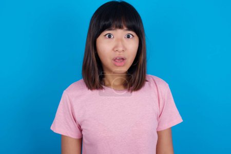 Photo for Emotional attractive young asian woman wearing t-shirt against blue background with opened mouth expresses great surprise and fright, stares at camera. Unexpected shocking news and human reaction. - Royalty Free Image