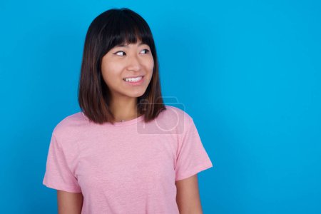Photo for Oops! Portrait of young asian woman wearing t-shirt against blue background clenches teeth and looks confusedly aside, realizes her bad mistake, - Royalty Free Image