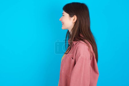 Photo for Profile of smiling young caucasian girl wearing pink shirt isolated over blue background with healthy skin, has contemplative expression, ready to have outdoor walk. - Royalty Free Image