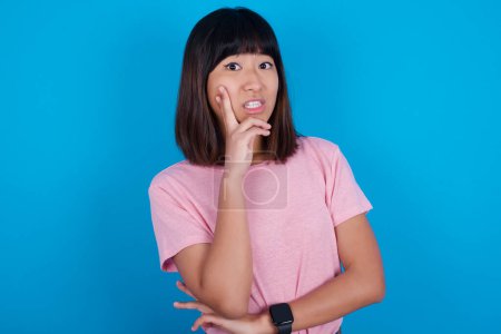 Téléchargez les photos : Jeune femme asiatique portant t-shirt sur fond bleu couvrant la bouche avec les mains effrayées de quelque chose ou quelqu'un mordant ongles - en image libre de droit
