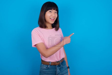 Téléchargez les photos : Jeune femme asiatique portant t-shirt sur fond bleu pointant du doigt et vous souriant. Regarde là-bas.! - en image libre de droit
