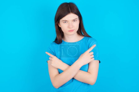 Foto de Chica caucásica joven seria con camiseta azul aislada sobre fondo azul cruza las manos y puntos en diferentes lados duda entre dos artículos. Concepto de decisión difícil - Imagen libre de derechos