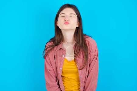 Photo for Young caucasian girl wearing pink shirt isolated over blue background keeps lips as going to kiss someone, has glad expression, grimace face. Standing indoors. Beauty concept. - Royalty Free Image