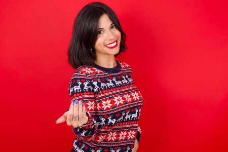 Téléchargez les photos : Brune femme caucasienne portant un pull de Noël sur fond rouge invitant à venir avec la main. Heureux que tu sois venu - en image libre de droit