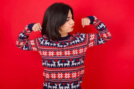 Téléchargez les photos : Brune caucasienne portant un pull de Noël sur fond rouge montrant les muscles des bras souriant fier. Concept de fitness. - en image libre de droit
