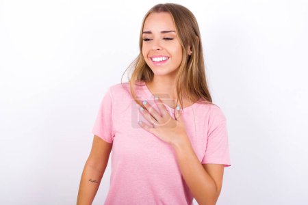 Joyful Young Caucasian girl wearing pink T-shirt on white background expresses positive emotions recalls something funny keeps hand on chest and giggles happily.