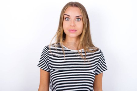 Stunned beautiful blonde girl wearing striped t-shirt on white background stares reacts on shocking news. Astonished MODEL holds breath