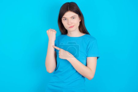Téléchargez les photos : Jeune fille caucasienne portant bleu T-shirt isolé sur fond bleu à la hâte pointant vers regarder le temps, impatience, bouleversé et en colère pour retard de délai - en image libre de droit
