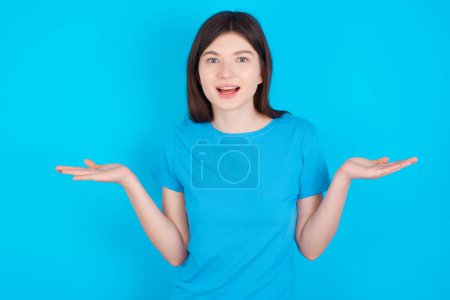 Foto de ¿Y qué? Retrato de una joven arrogante caucásica usando una camiseta azul aislada sobre fondo azul encogiendo las manos de lado sonriendo jadeando indiferente, diciendo algo obvio. - Imagen libre de derechos