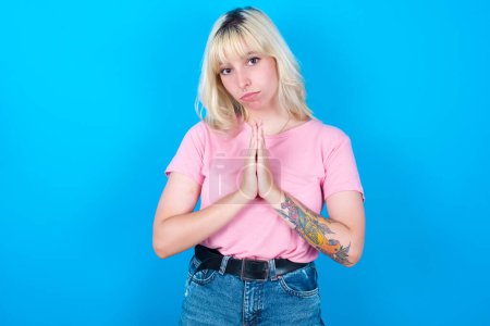 Photo for Caucasian girl wearing pink t-shirt isolated over blue background keeps palms pressed together in front of her having regretful look, asking for forgiveness. Forgive me please. - Royalty Free Image