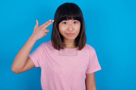 Téléchargez les photos : Malheureux jeune asiatique femme porter t-shirt sur bleu fond imite pistolet tirer fait suicide geste garde deux doigts sur temples. - en image libre de droit