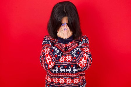 Téléchargez les photos : Brune caucasienne portant un pull de Noël sur fond rouge couvrant son visage de ses mains, étant dévastée et pleurant. Triste concept - en image libre de droit