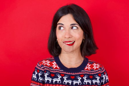 Téléchargez les photos : Incroyable brune caucasienne portant un pull de Noël sur fond rouge lèvre mordante et regardant délicat à vide espace. - en image libre de droit