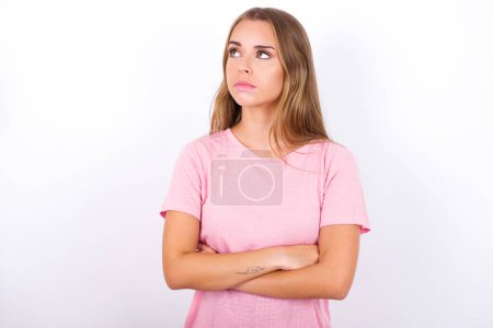 Foto de Encantadora y pensativa joven caucásica con camiseta rosa sobre fondo blanco se levanta con los brazos cruzados concentrado en algún lugar con expresión pensativa piensa qué hacer - Imagen libre de derechos