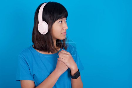 Téléchargez les photos : Jeune femme asiatique portant t-shirt sur fond bleu porte casque stéréo écouter de la musique concentrée et regarder de côté avec intérêt. - en image libre de droit