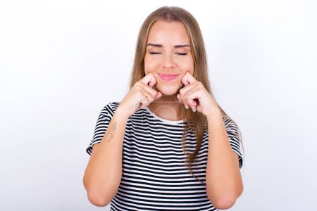 Pleased beautiful blonde girl wearing striped t-shirt on white background with closed eyes keeps hands near cheeks and smiles tenderly imagines something very pleasant
