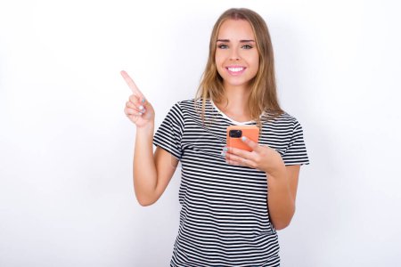 Foto de Sonriente hermosa chica rubia con una camiseta a rayas sobre fondo blanco apuntando con el dedo al espacio en blanco sosteniendo el teléfono en una mano - Imagen libre de derechos