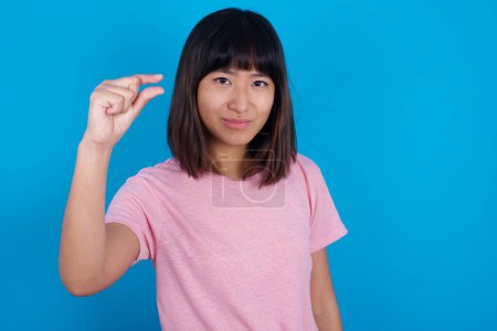Photo for Upset young asian woman wearing t-shirt against blue background shapes little gesture with hand demonstrates something very tiny small size. Not very much - Royalty Free Image