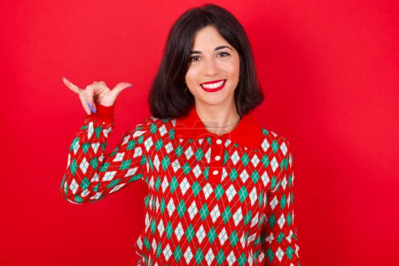 Téléchargez les photos : Brunette femme caucasienne portant chandail de Noël sur fond rouge montrant le numéro six Liu avec des doigts geste en langue chinoise des signes - en image libre de droit