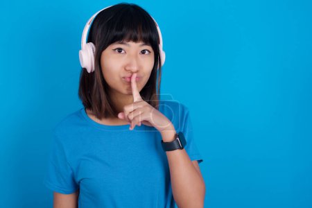 Photo for Young asian woman wearing t-shirt against blue background making hush gesture with finger on her lips wearing  wireless headphones. Be quiet. - Royalty Free Image