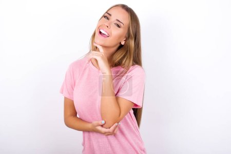 Photo for Young Caucasian girl wearing pink T-shirt on white background laughs happily keeps hand on chin expresses positive emotions smiles broadly has carefree expression - Royalty Free Image