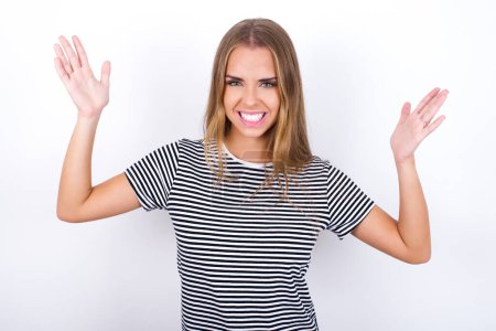 Crazy outraged beautiful blonde girl wearing striped t-shirt on white background screams loudly and gestures angrily yells furiously. Negative human emotions feelings concept