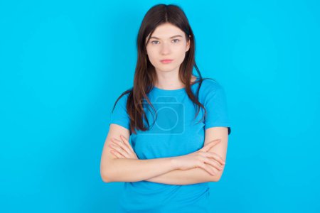 Photo for Confident young caucasian girl wearing blue T-shirt isolated over blue background with arms crossed looking to the camera - Royalty Free Image