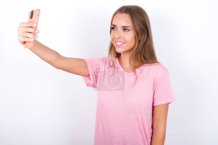 Isolated shot of pleased cheerful Young Caucasian girl wearing pink T-shirt on white background makes selfie with mobile phone. People, technology and leisure concept