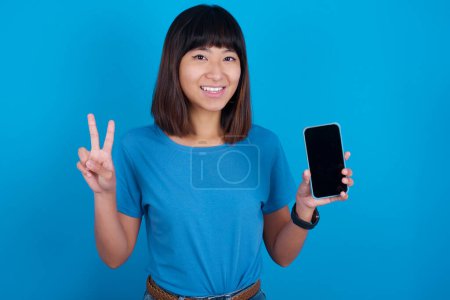 Photo for Young asian woman wearing t-shirt against blue background holding modern device showing v-sign - Royalty Free Image