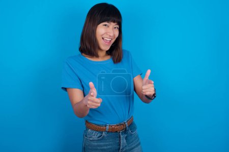 Téléchargez les photos : Jeune femme asiatique portant t-shirt sur fond bleu dirige les doigts à la caméra sélectionne quelqu'un. Je vous recommande. Meilleur choix - en image libre de droit