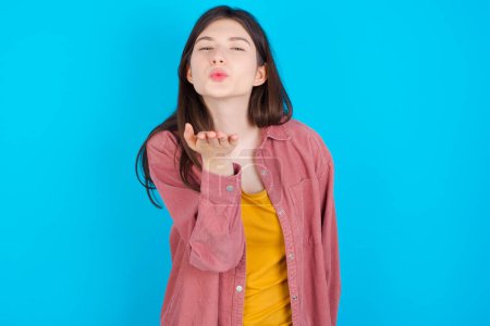 Téléchargez les photos : Jeune fille caucasienne portant chemise rose isolé sur fond bleu regardant la caméra souffler un baiser avec la main sur l'air étant belle et sexy. Expression d'amour. - en image libre de droit
